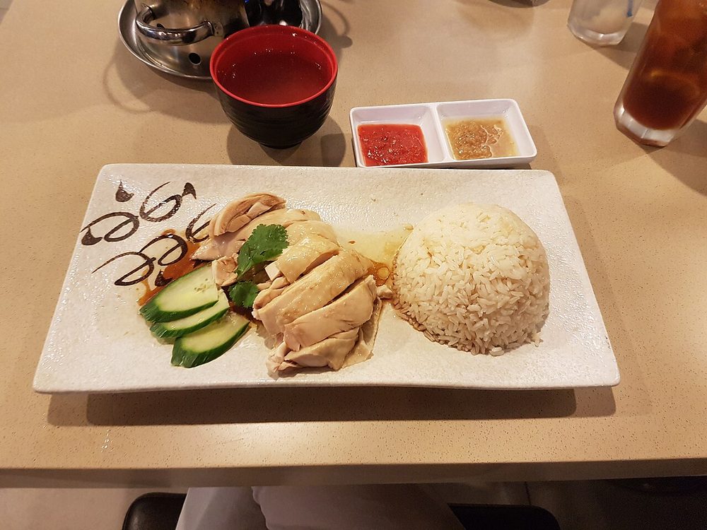 A white rectangular plate of sliced poached chicken with cucumber and a moulded mound of rice, with a two-compartment dish of chilli and ginger sauce and a bowl of soup behind