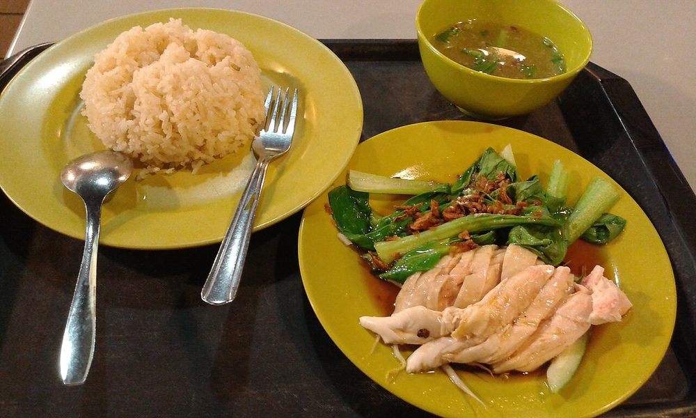 A plate of sliced poached white chicken with greens and garnish beside a dome of rice and a small bowl of soup, on a hawker centre tray in Singapore