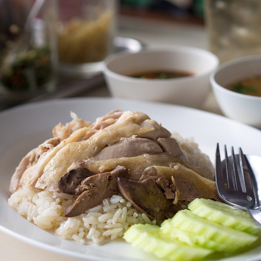 Thai khao man kai: sliced poached chicken and liver on rice with cucumber, with bowls of broth and sauce behind