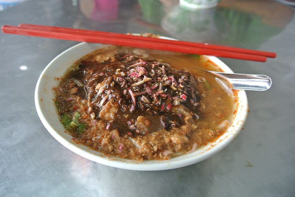 A bowl of asam laksa at Air Itam market in Penang, topped with flaked fish, shredded vegetables and a dark smear of prawn paste, with red chopsticks