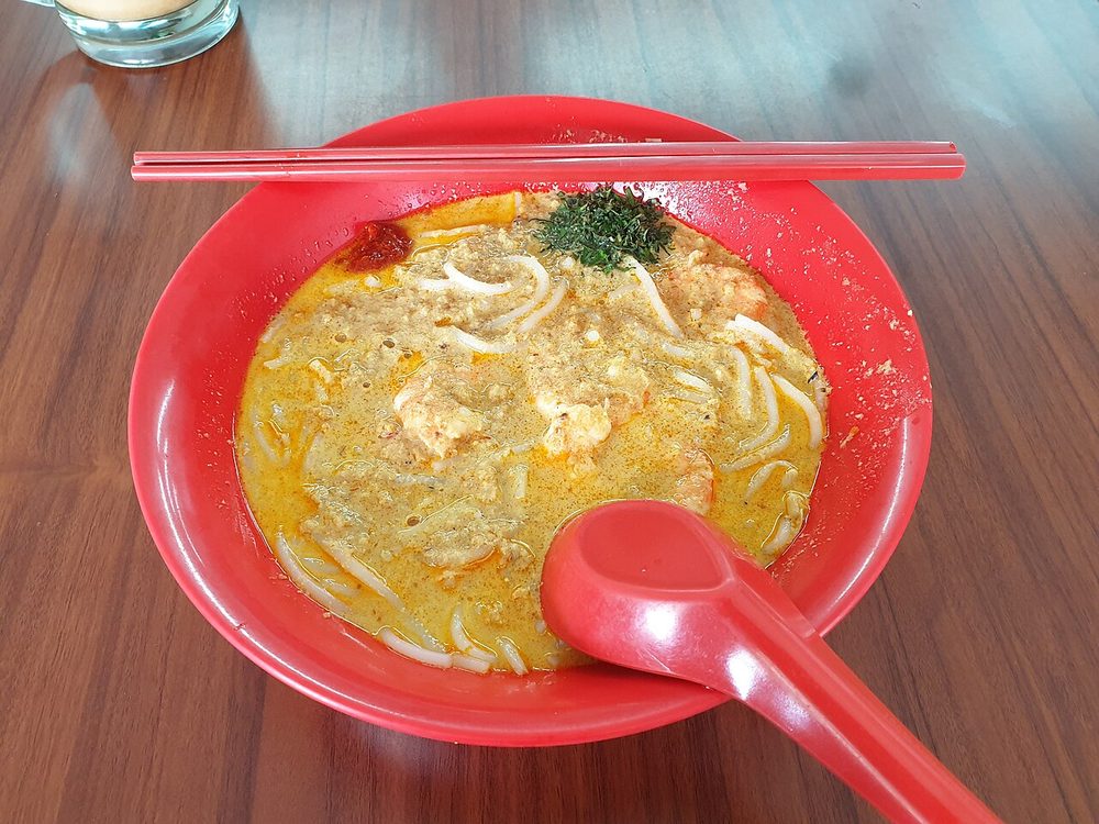 A red bowl of Katong laksa seen from above: thick rice noodles in orange coconut gravy, with chopped laksa leaf, a red plastic spoon and chopsticks on a wooden table
