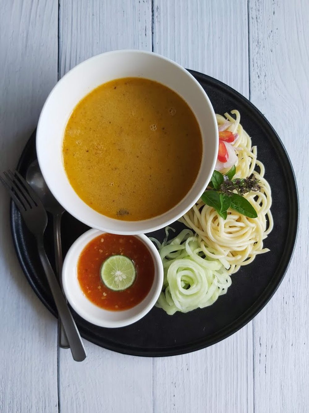 Laksa Johor seen from above: spaghetti on a black plate with cucumber and herbs, a bowl of thick orange fish gravy, and a saucer of sambal with a calamansi half