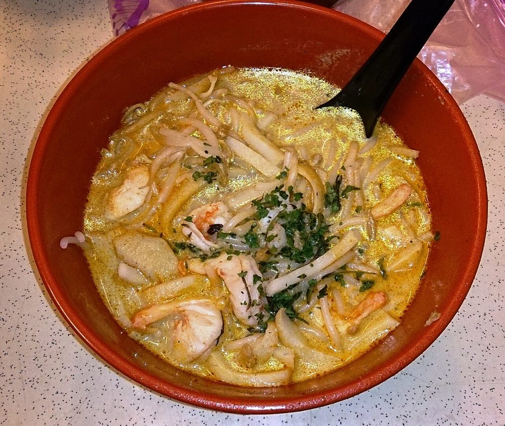 A close-up of Katong laksa in a red bowl: short-cut rice noodles, prawns, cockles and fishcake in orange gravy, scattered with chopped laksa leaf, with a black plastic spoon
