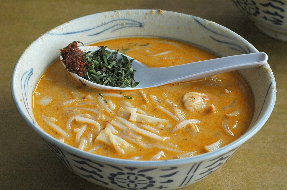 A blue and white bowl of Katong laksa with a porcelain spoon resting in it, holding chopped laksa leaf and sambal
