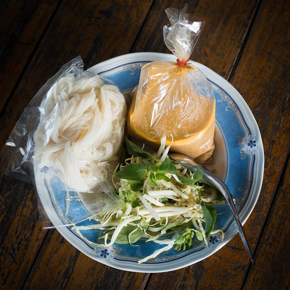 Khanom chin nam ya from a Thai market: coils of white rice noodles, a plastic bag of orange fish curry and a pile of fresh bean sprouts and herbs on a blue plate