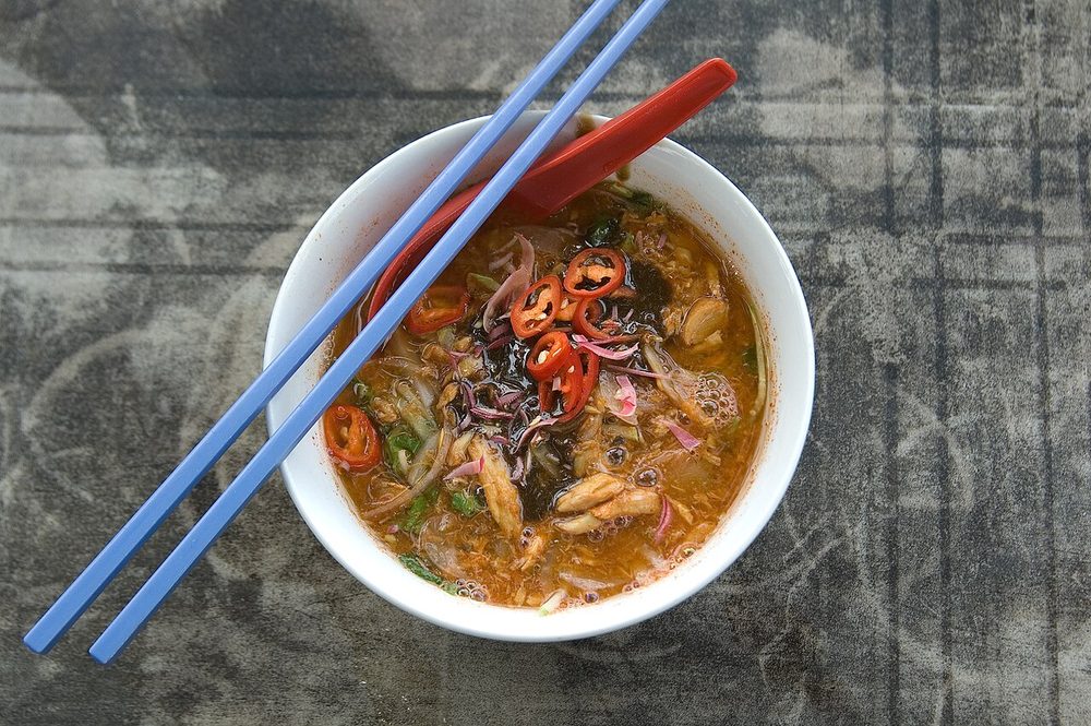 A white bowl of Penang asam laksa seen from above, with sliced red chilli, onion, mint and cucumber on a dark broth, and blue chopsticks