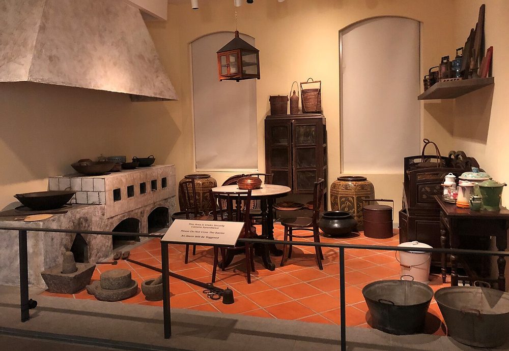 A reconstructed interwar Peranakan kitchen in a museum, with a tiled wood-fired stove, stone mortars, clay pots and wooden cupboards
