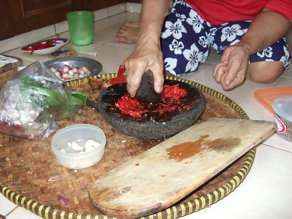 A woman pounding red chilli paste in a stone mortar on a woven tray, with shallots and garlic beside her, in a home kitchen