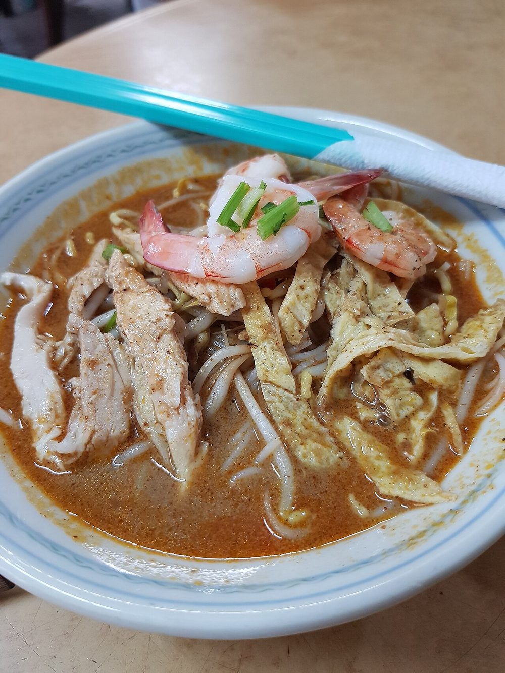 A bowl of Sarawak laksa with halved prawns, shredded chicken, omelette strips and bean sprouts in a brown-orange broth